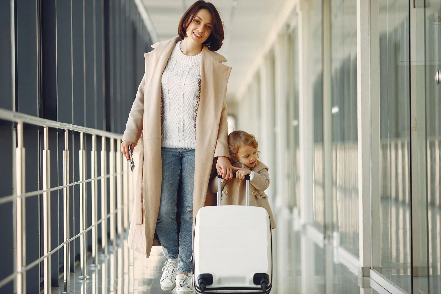 Mutter und kleine Tochter gehen mit Rollkoffer durch einen verglasten Flughafengang